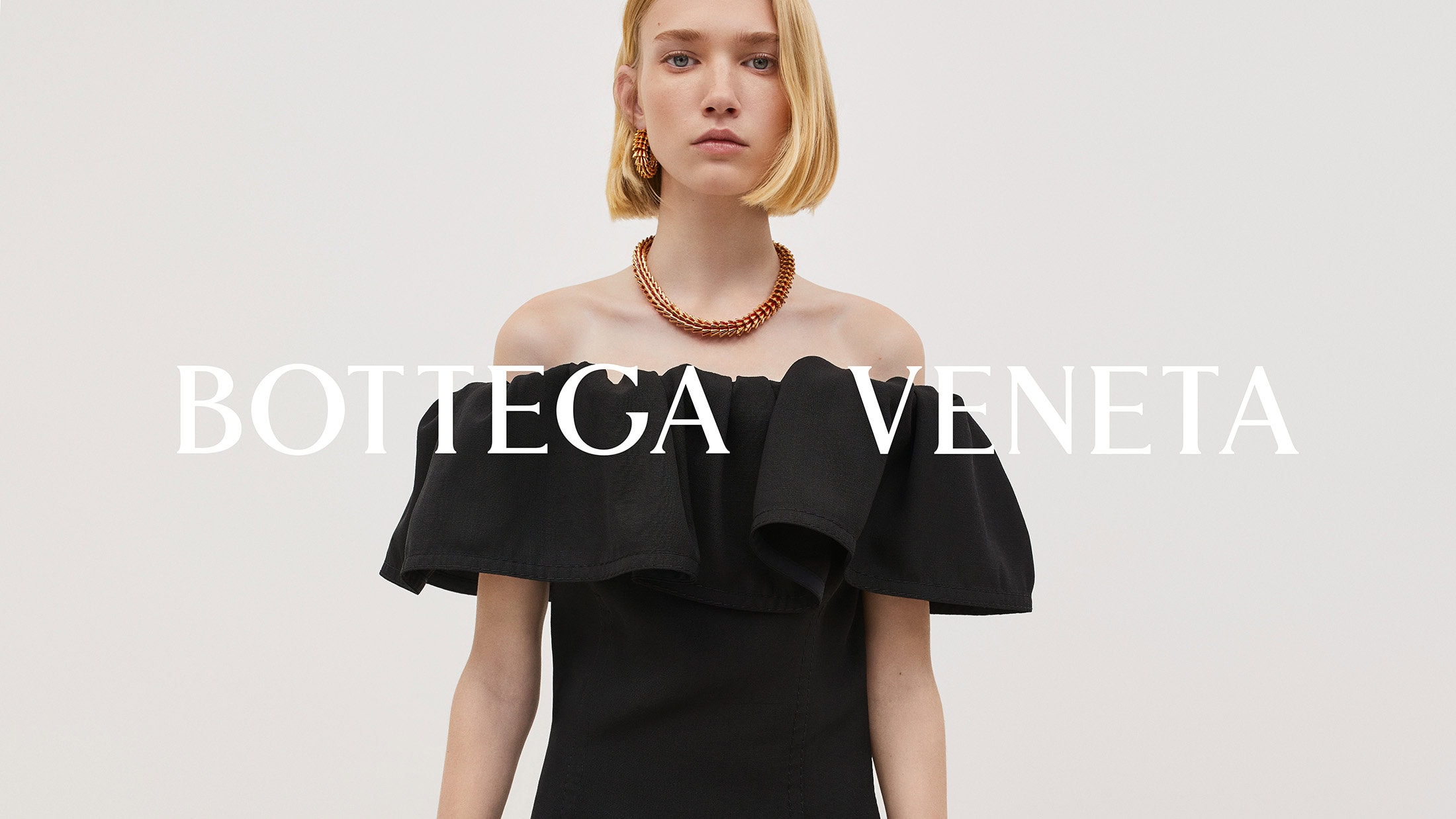 Elegant woman with blonde bob wearing an off-the-shoulder black dress and copper jewelry in a bright, minimalist studio setting.