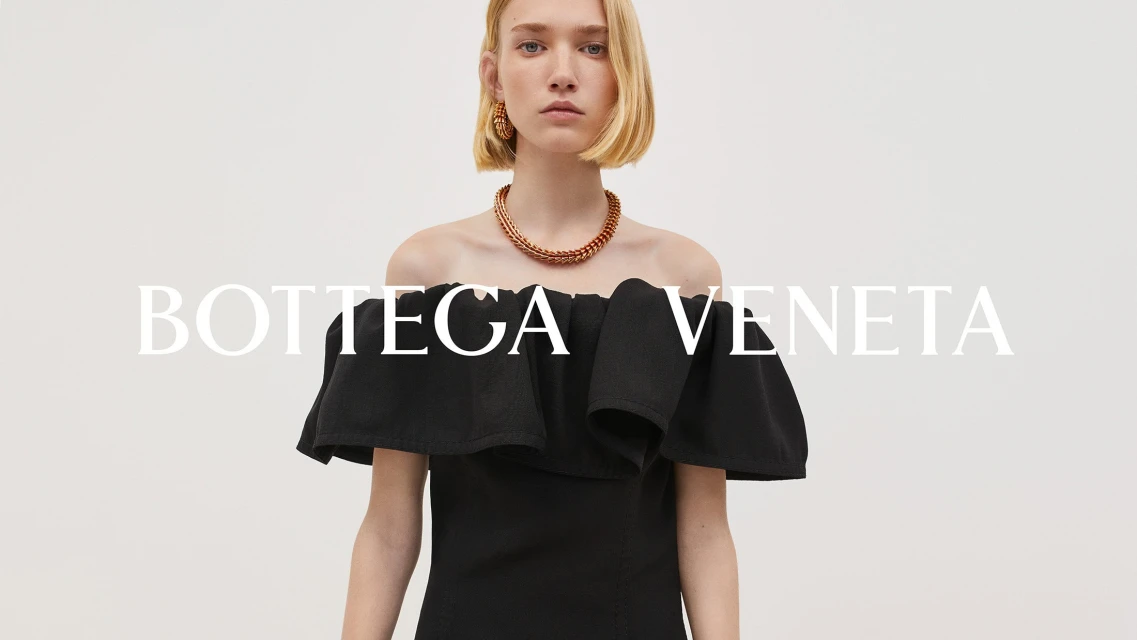 Elegant woman with blonde bob wearing an off-the-shoulder black dress and copper jewelry in a bright, minimalist studio setting.