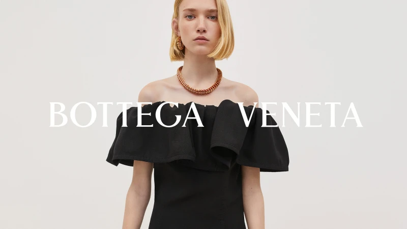 Elegant woman with blonde bob wearing an off-the-shoulder black dress and copper jewelry in a bright, minimalist studio setting.