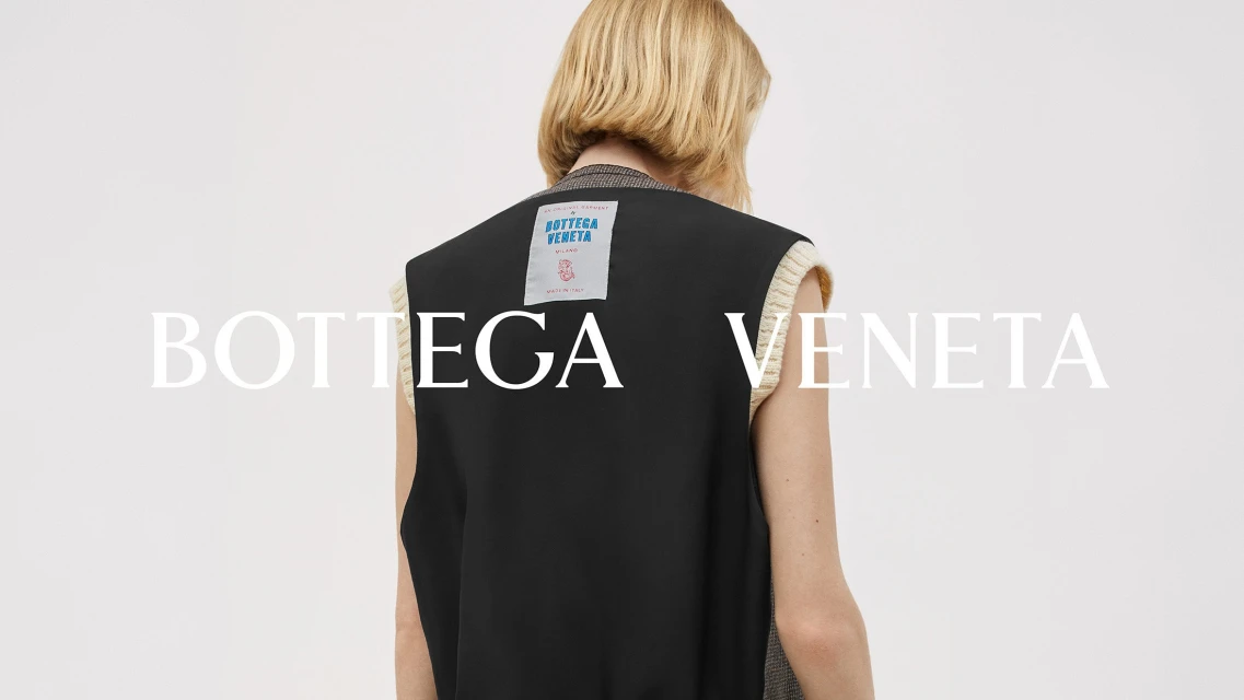Fashion shot of a woman wearing a black vest with cream trim, posing in front of a neutral background, with a Bottega Veneta logo.