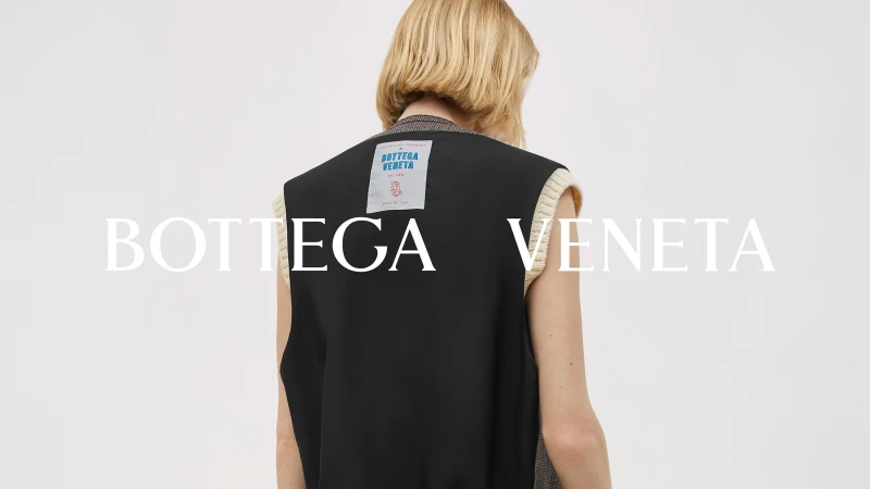 Fashion shot of a woman wearing a black vest with cream trim, posing in front of a neutral background, with a Bottega Veneta logo.
