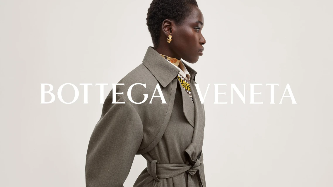 Profile shot of an adult African-American female fashion model wearing a gray coat and gold earrings set against a bright neutral background.