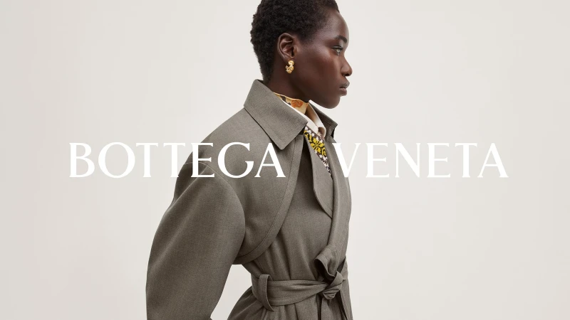 Profile shot of an adult African-American female fashion model wearing a gray coat and gold earrings set against a bright neutral background.
