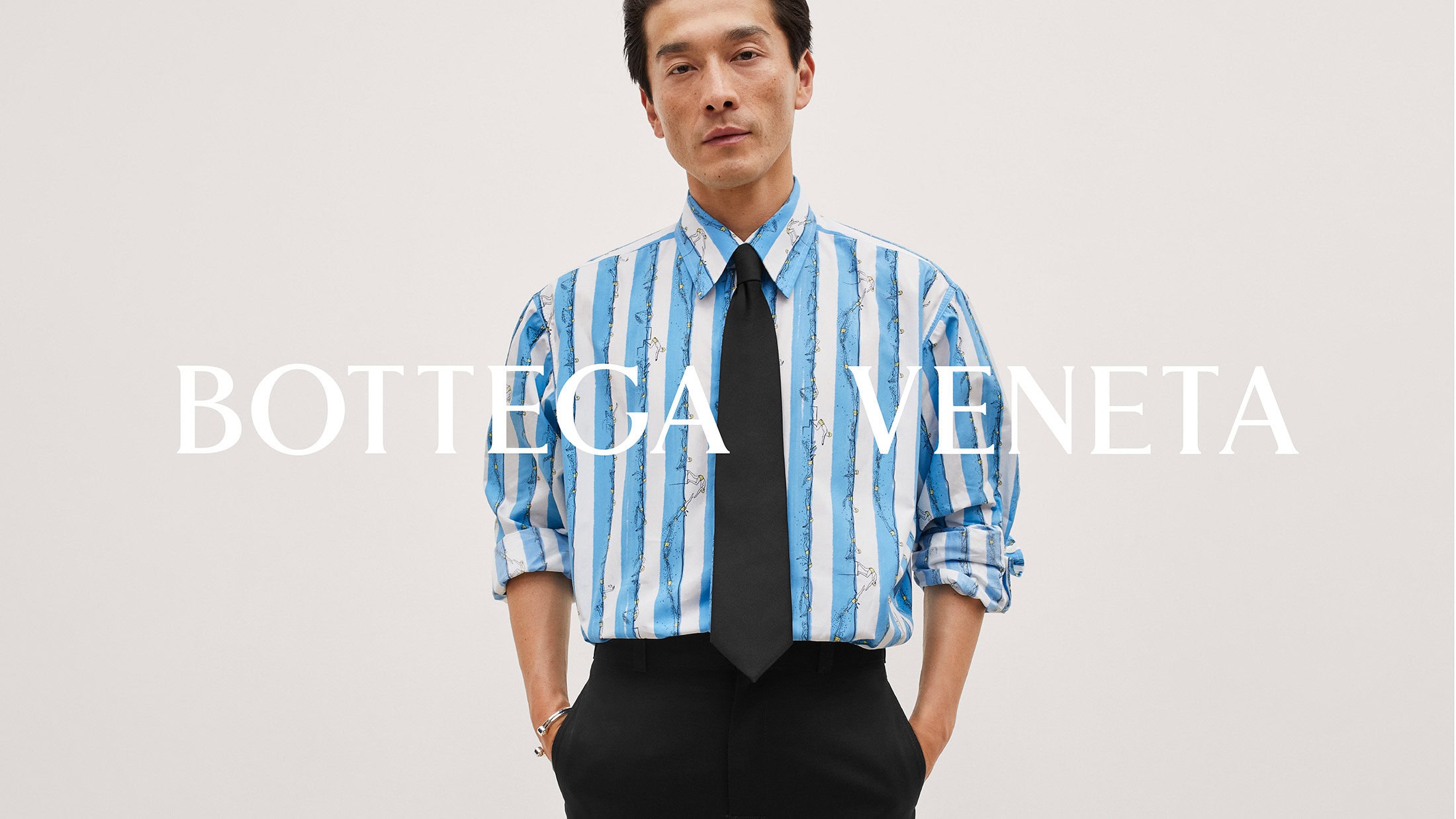 An Asian adult male model poses in a light blue striped shirt with a black tie and black trousers against a plain neutral background.