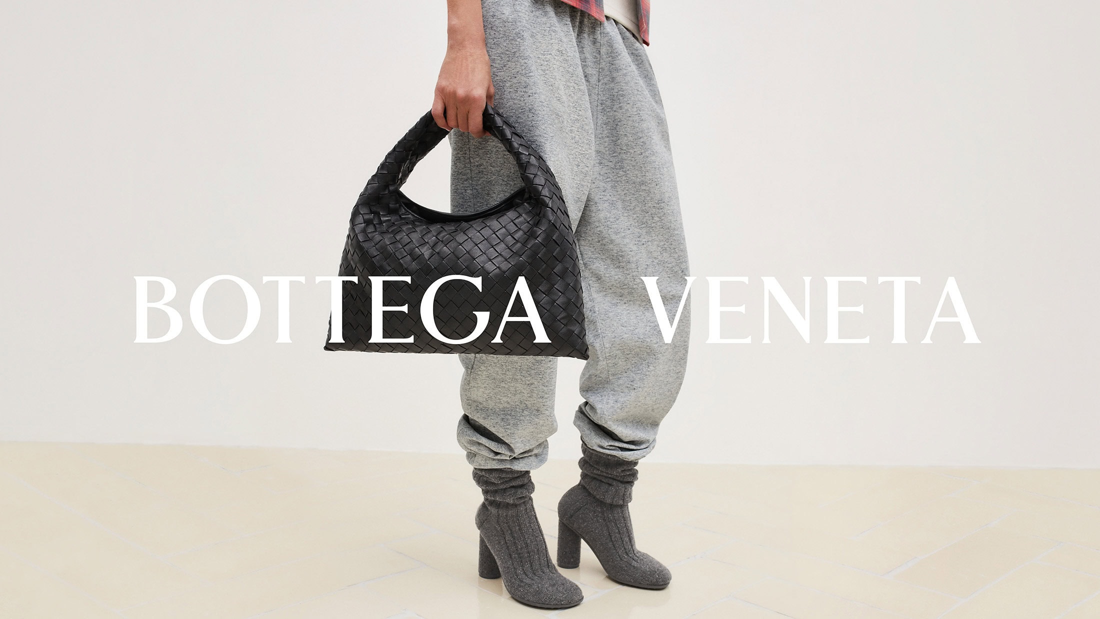 Fashion shot features a person wearing gray sweatpants and gray high heel boots, holding a black Bottega Veneta handbag against a white background.
