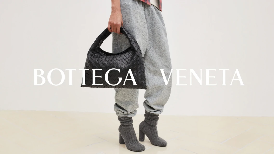 Fashion shot features a person wearing gray sweatpants and gray high heel boots, holding a black Bottega Veneta handbag against a white background.