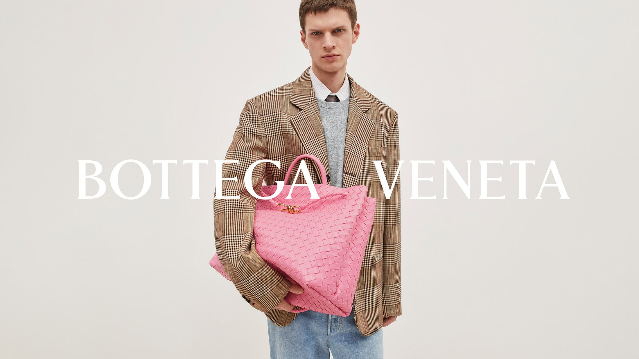 An adult male poses with a large, pink woven leather tote bag in a studio setting. He wears a plaid blazer and jeans, with 'Bottega Veneta' in large text.
