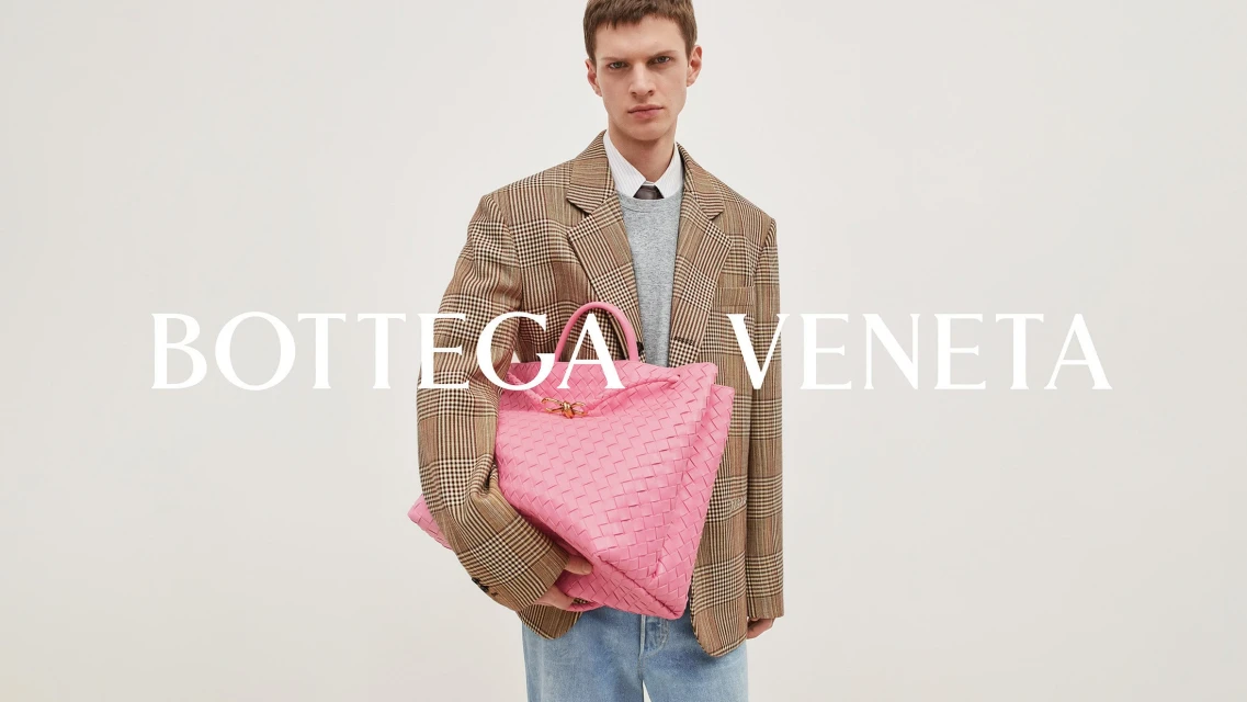 An adult male poses with a large, pink woven leather tote bag in a studio setting. He wears a plaid blazer and jeans, with 'Bottega Veneta' in large text.