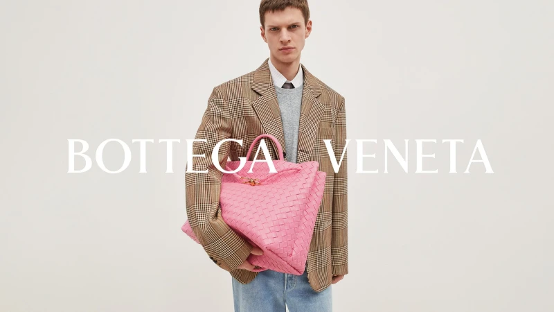 An adult male poses with a large, pink woven leather tote bag in a studio setting. He wears a plaid blazer and jeans, with 'Bottega Veneta' in large text.