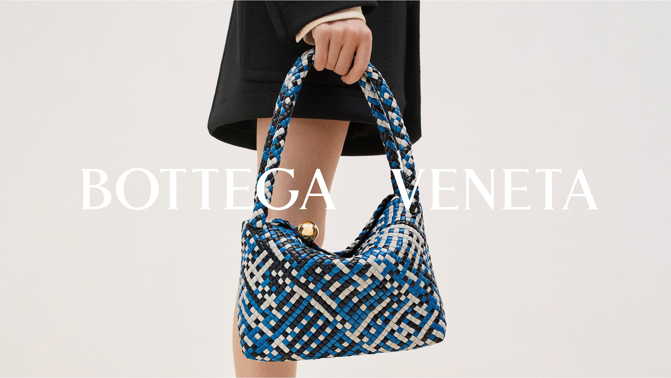 Woman holding a blue, white, and black woven Bottega Veneta bag with a gold clasp against a plain white background in a studio setting.