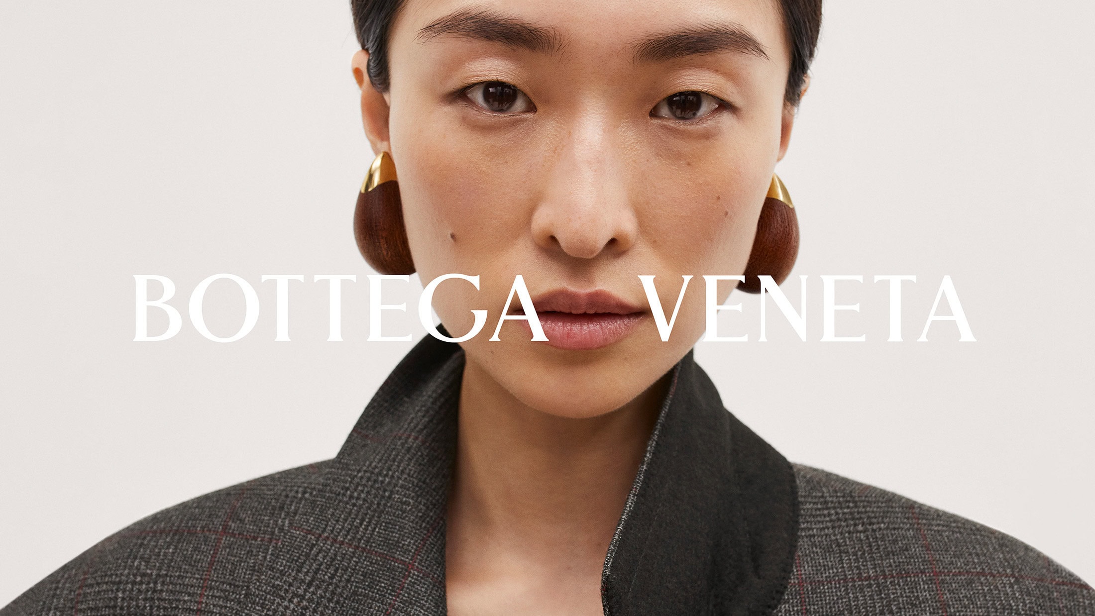 Close-up of an Asian woman with gold and brown earrings, wearing a grey plaid blazer, against a pale background, with 'BOTTEGA VENETA' in white text.