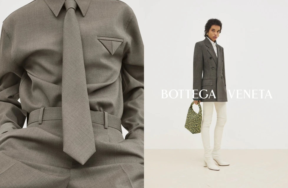 Fashion editorial featuring an adult female model wearing a grey tailored shirt, tie, blazer, and cream-colored pants in a studio.