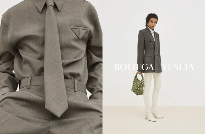 Fashion editorial featuring an adult female model wearing a grey tailored shirt, tie, blazer, and cream-colored pants in a studio.
