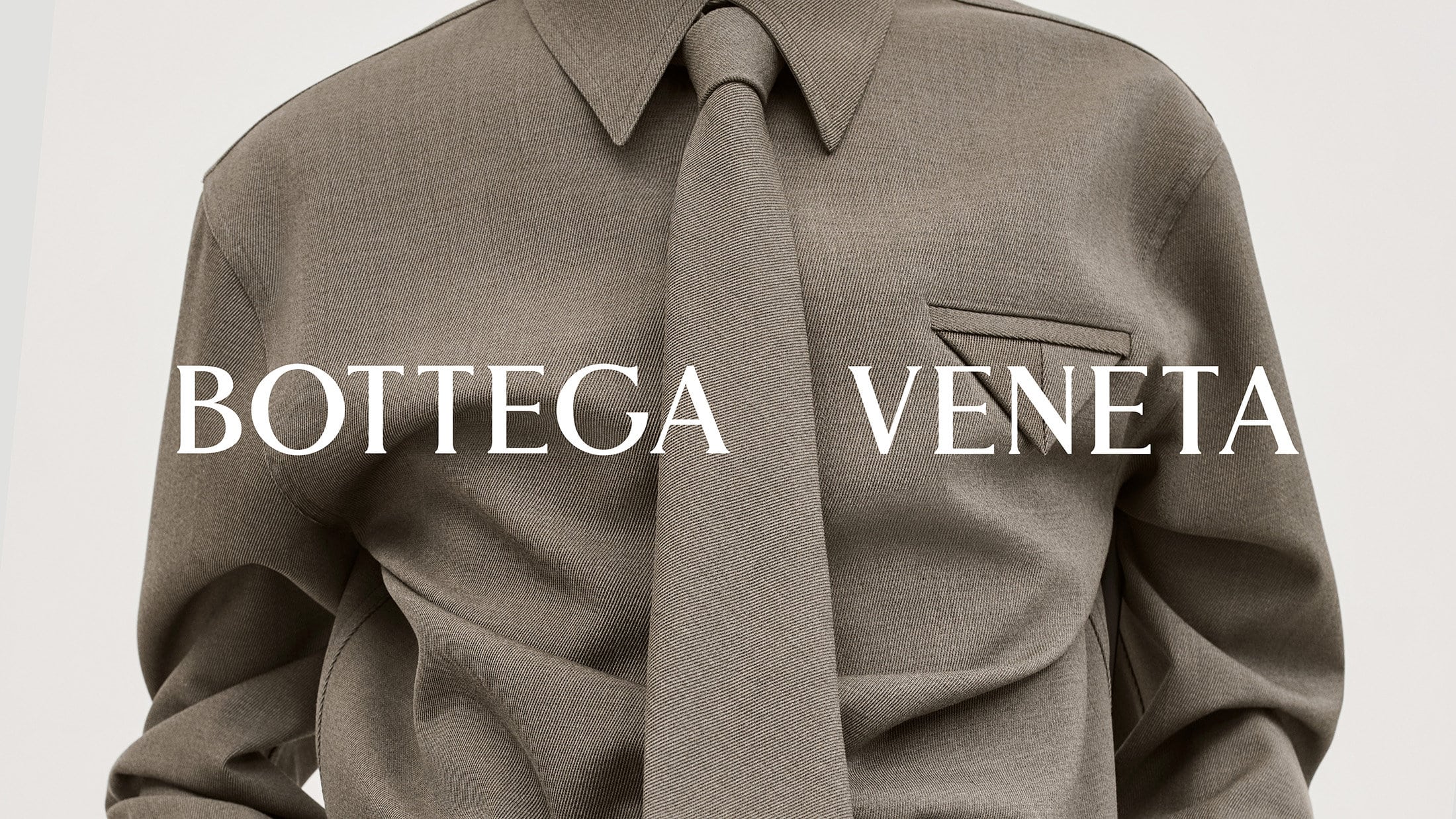 Close-up of a person wearing a grey collared shirt and tie, featuring the 'Bottega Veneta' logo, studio lit on a neutral backdrop.