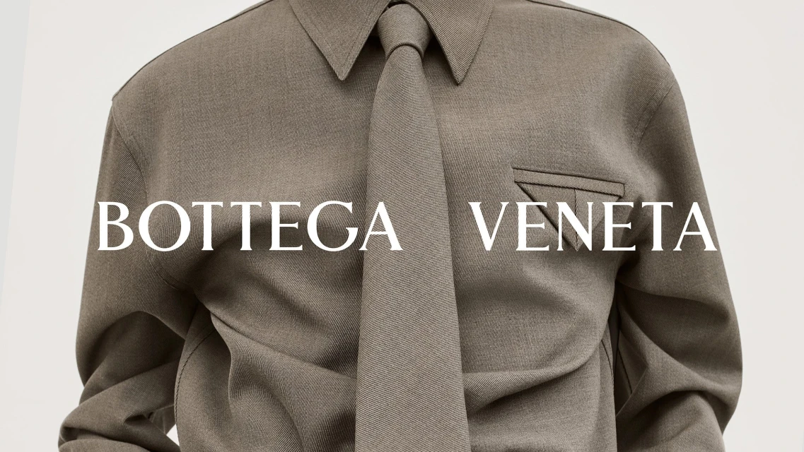 Close-up of a person wearing a grey collared shirt and tie, featuring the 'Bottega Veneta' logo, studio lit on a neutral backdrop.
