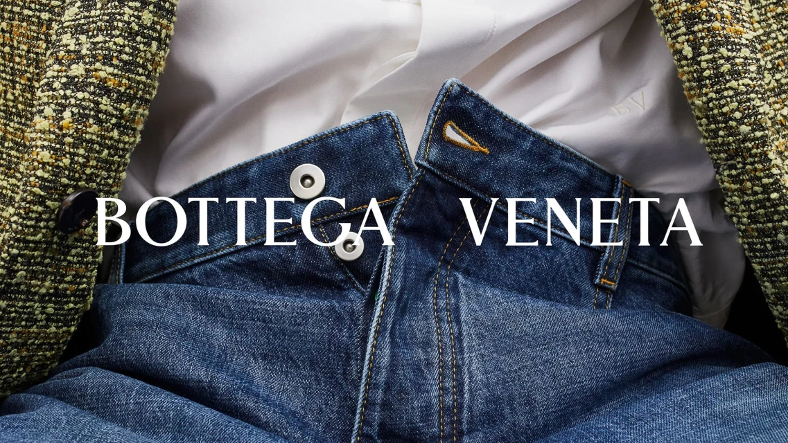 Close-up fashion shot: blue jeans paired with a white button-down shirt, layered under a tweed jacket, featuring Bottega Veneta logo.