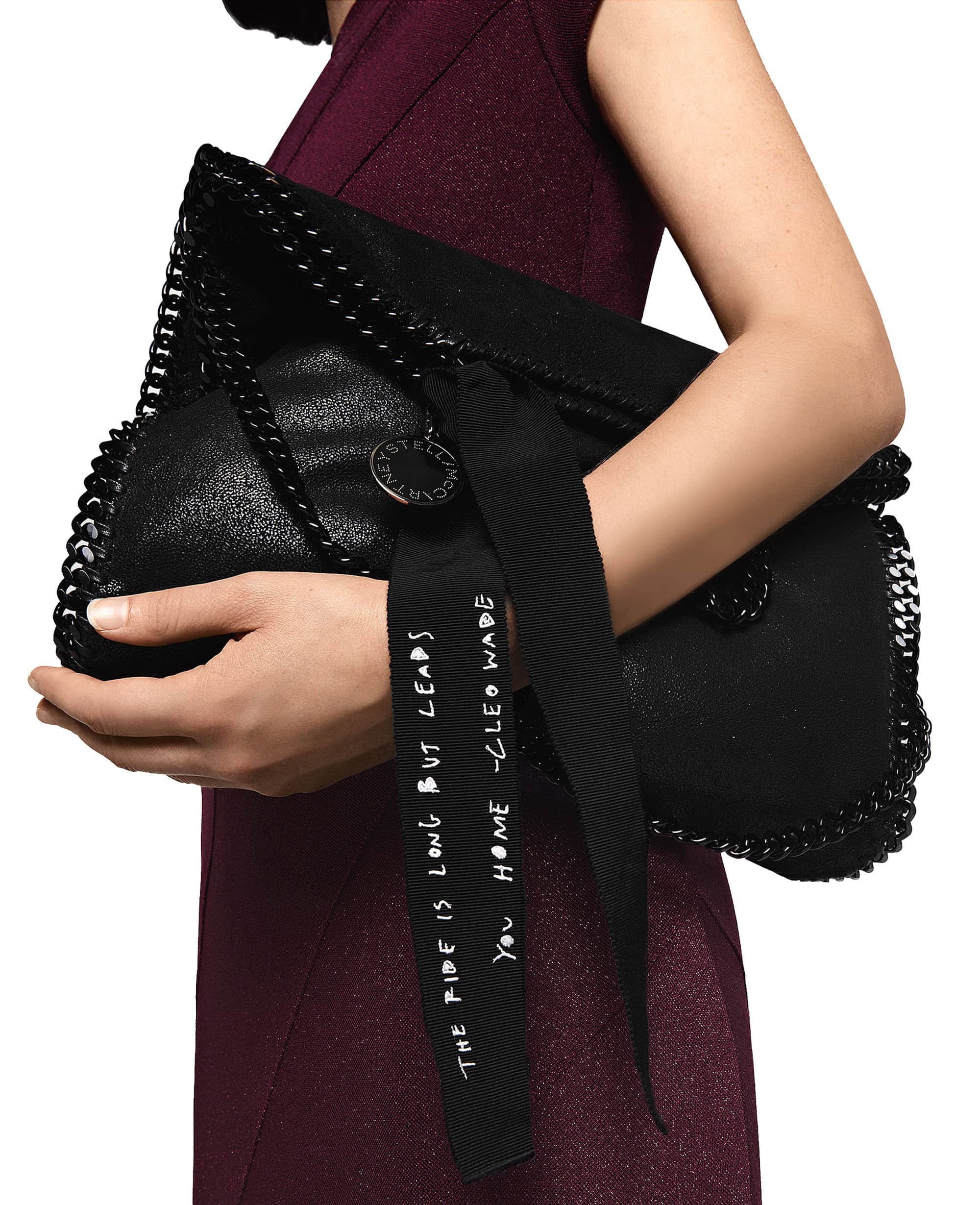 Black Stella McCartney clutch bag held by a fair-skinned adult woman wearing a maroon dress against a white backdrop in a studio shot.