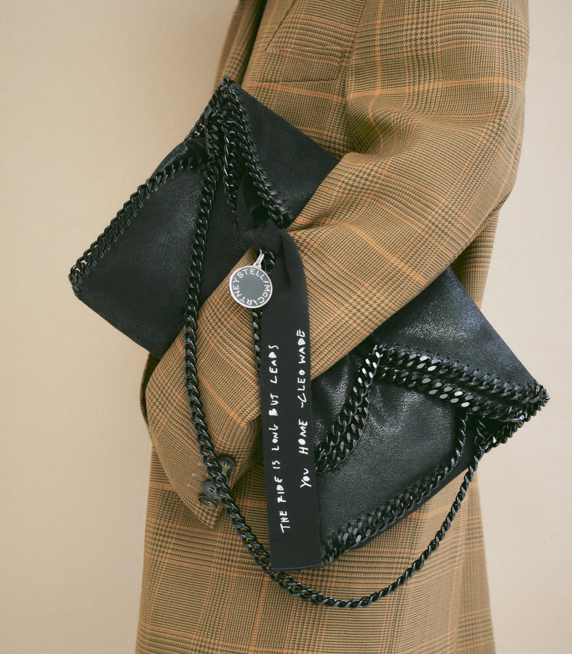 A black Stella McCartney clutch bag with chain detail and logo tag is held against a tan plaid blazer in a studio setting.