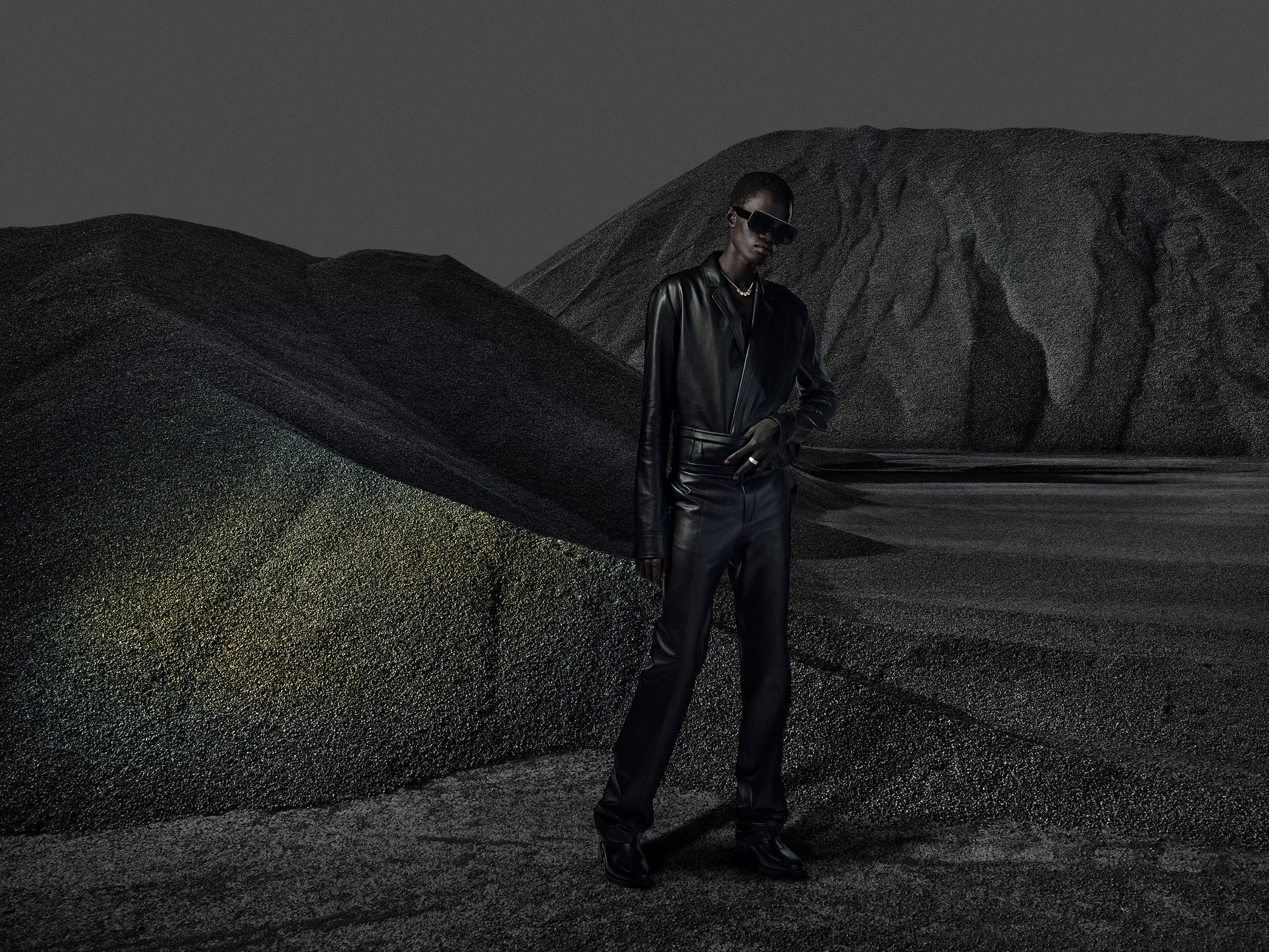 A person in a black leather outfit and sunglasses poses in a grayscale landscape with gravel and mountains, with desaturated colors.