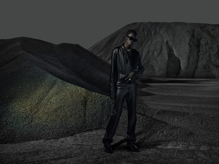 A person in a black leather outfit and sunglasses poses in a grayscale landscape with gravel and mountains, with desaturated colors.