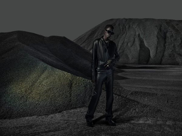A person in a black leather outfit and sunglasses poses in a grayscale landscape with gravel and mountains, with desaturated colors.