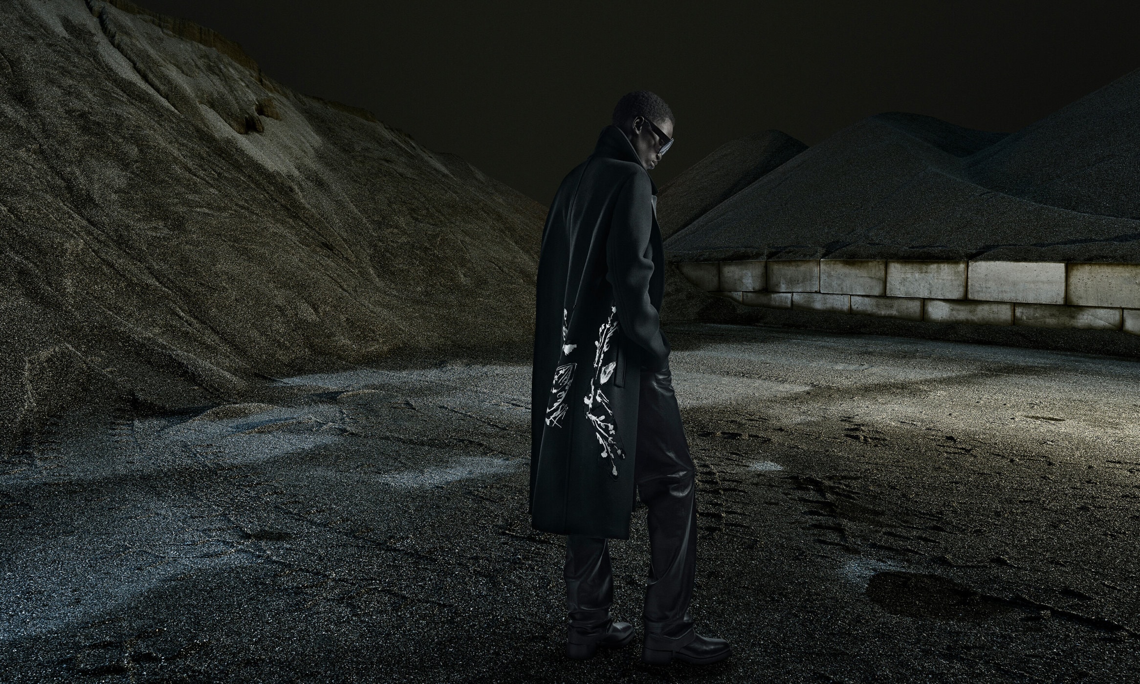 An adult male model in a dark coat with white detailing, standing against an industrial gravel-mountain background in low-key lighting.