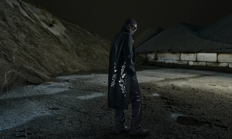 An adult male model in a dark coat with white detailing, standing against an industrial gravel-mountain background in low-key lighting.
