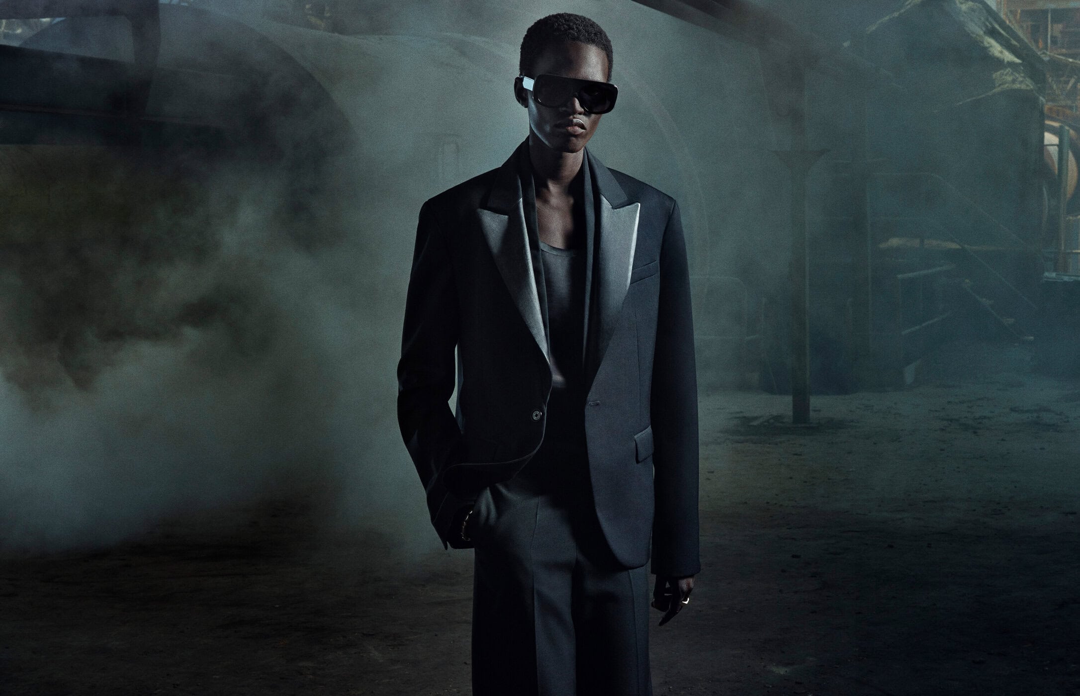 A person in a dark suit and sunglasses stands in a smoky, industrial setting, creating a mysterious and fashionable editorial image.