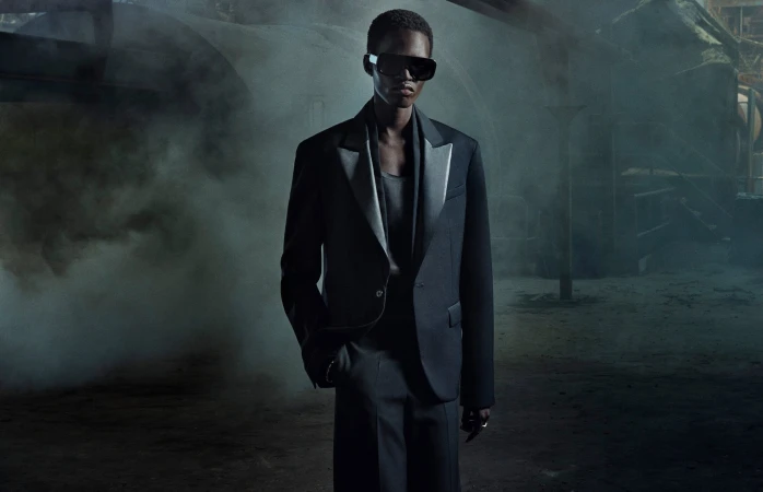 Mamuor Majeng poses in a tailored dark suit and black sunglasses amidst rising smoke inside an industrial facility.