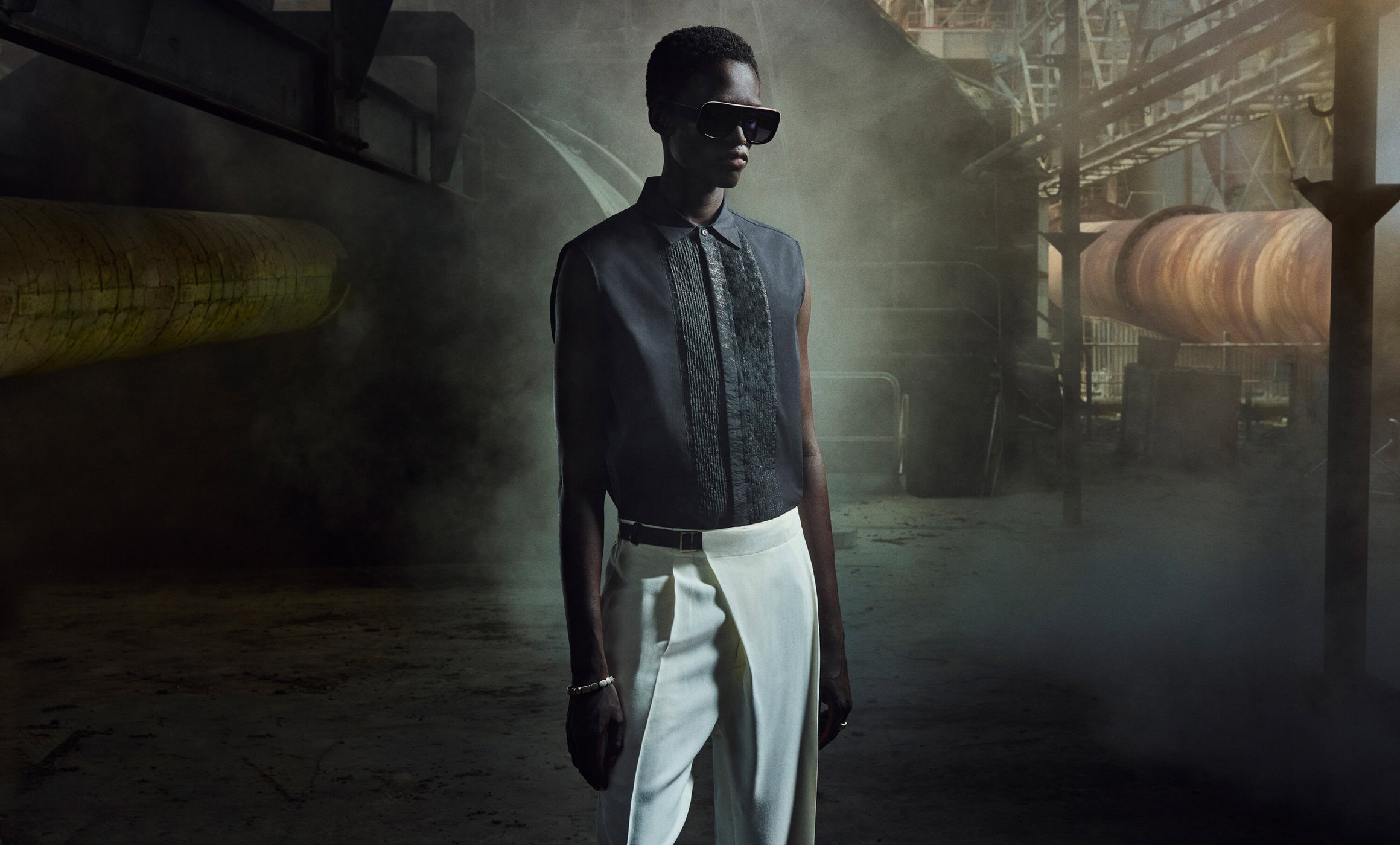 Fashion shot: a slender African-American man in black shirt and white pants stands in an industrial setting with pipes and machinery.