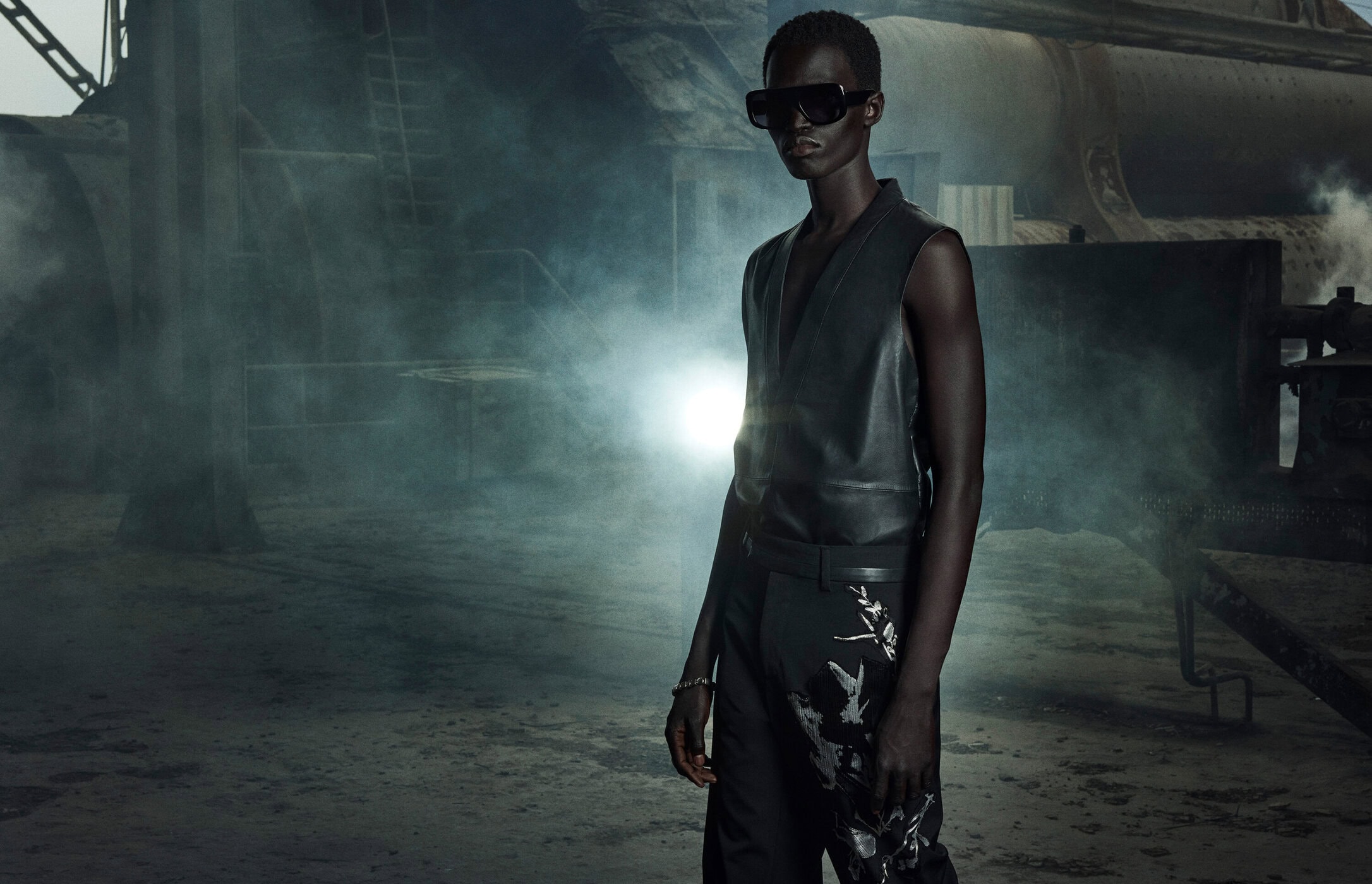 Fashion shot of an adult male model with dark skin and sunglasses wearing a vest and pants with floral prints in an industrial setting.