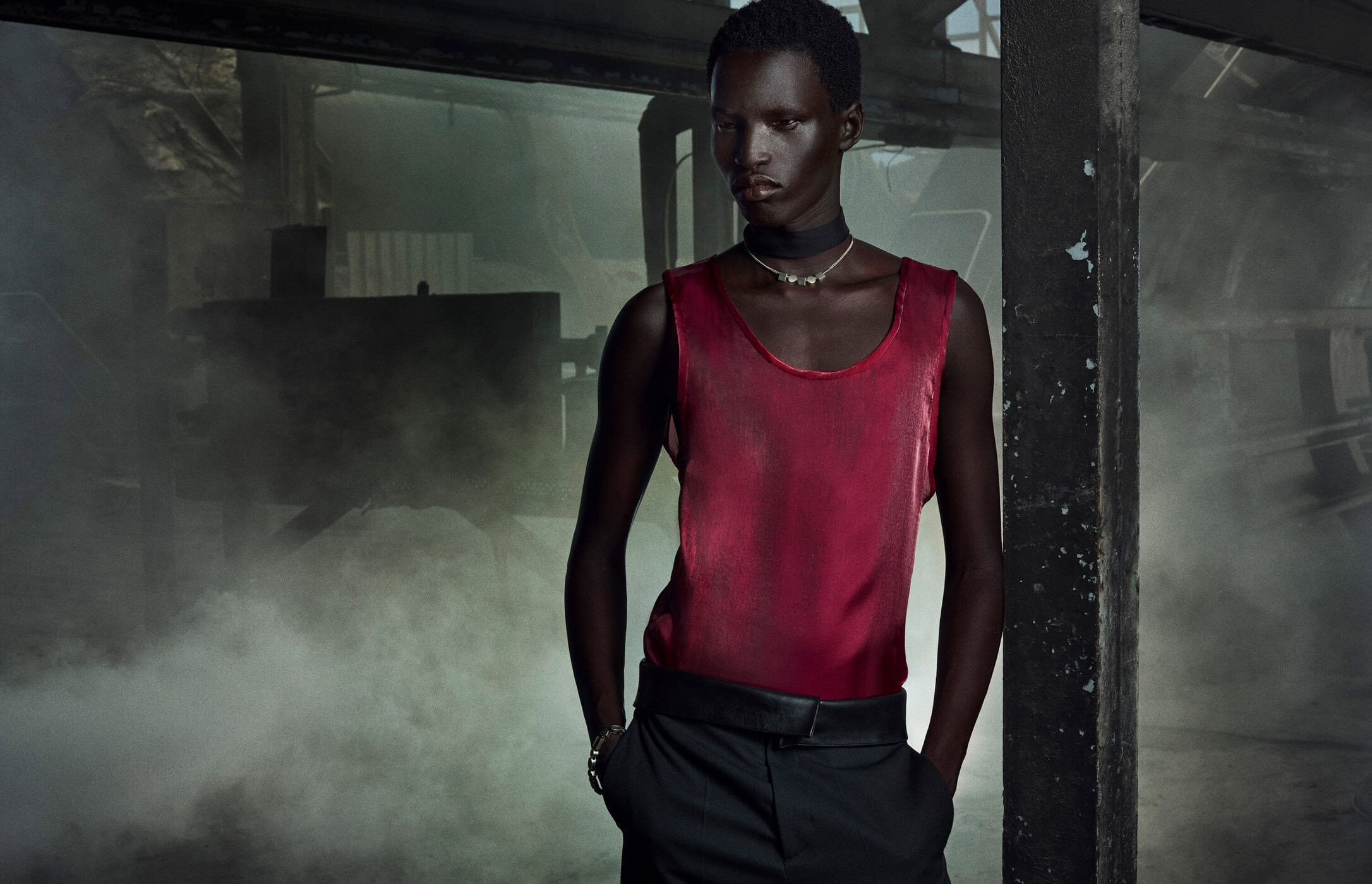 Fashion shot of a Black male model wearing a maroon tank top with black pants in an industrial setting with smoke and dark lighting.