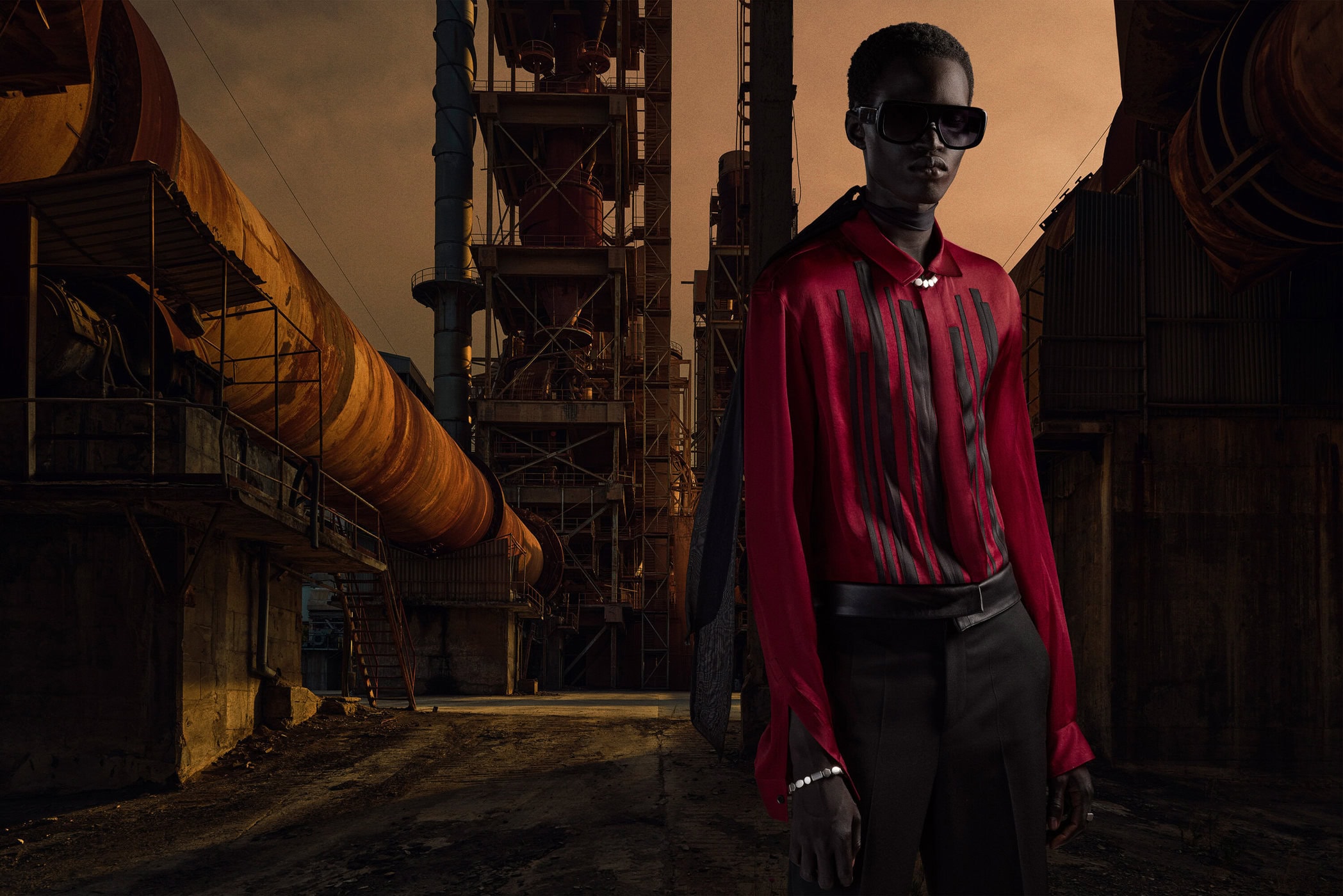 Fashion shot of a dark-skinned male model in a red shirt and black pants, posing in an industrial environment with metal structures and pipes.