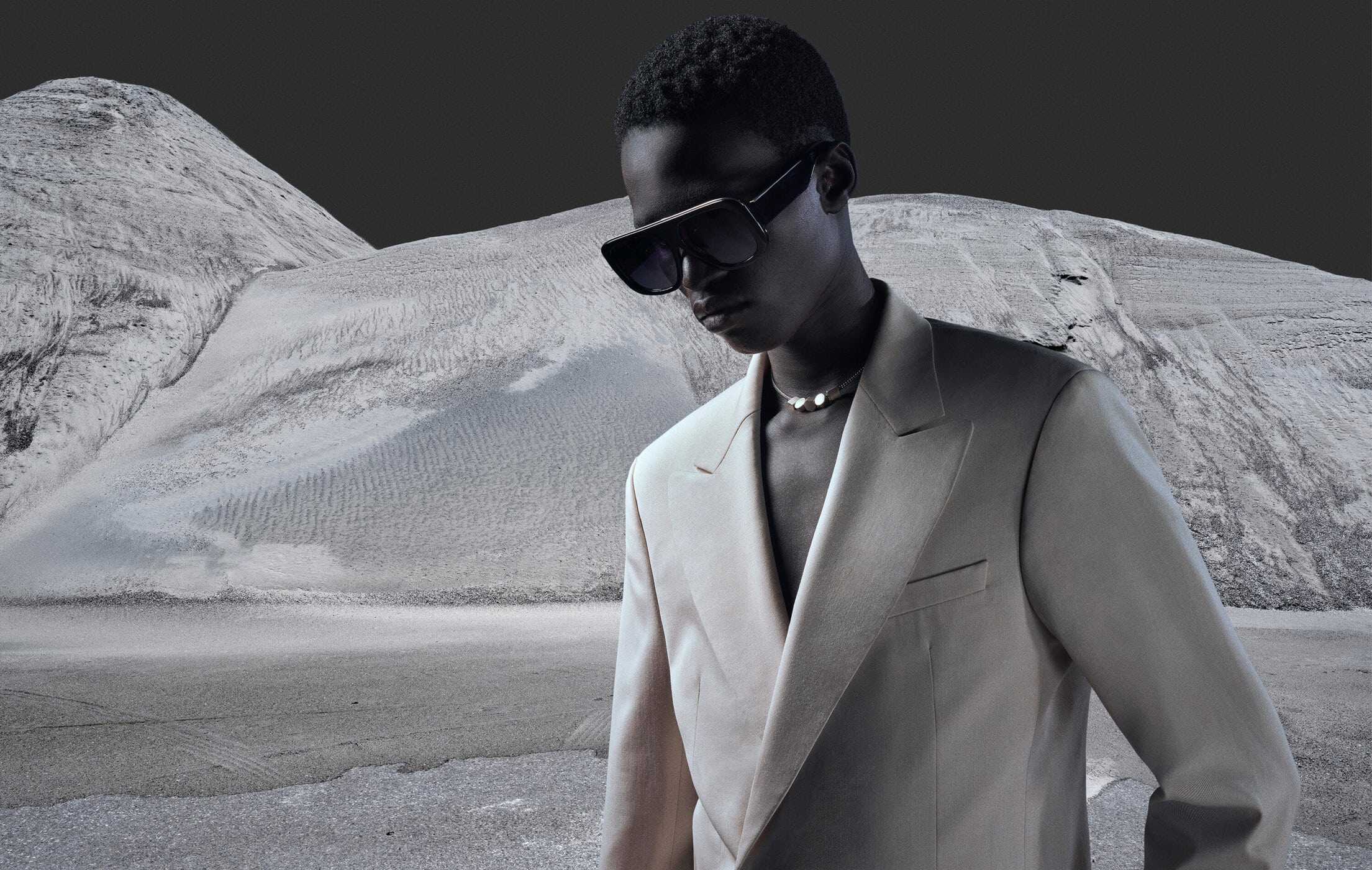 Fashion editorial shot of a Black male model in a cream suit and sunglasses standing against a stark desert landscape.