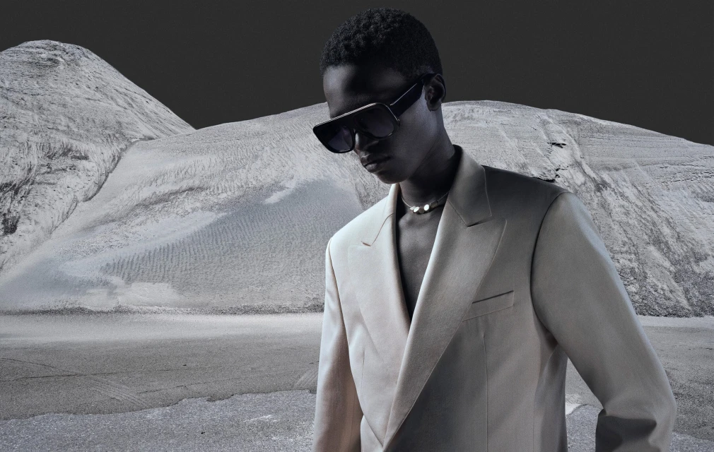 Fashion editorial shot of a Black male model in a cream suit and sunglasses standing against a stark desert landscape.