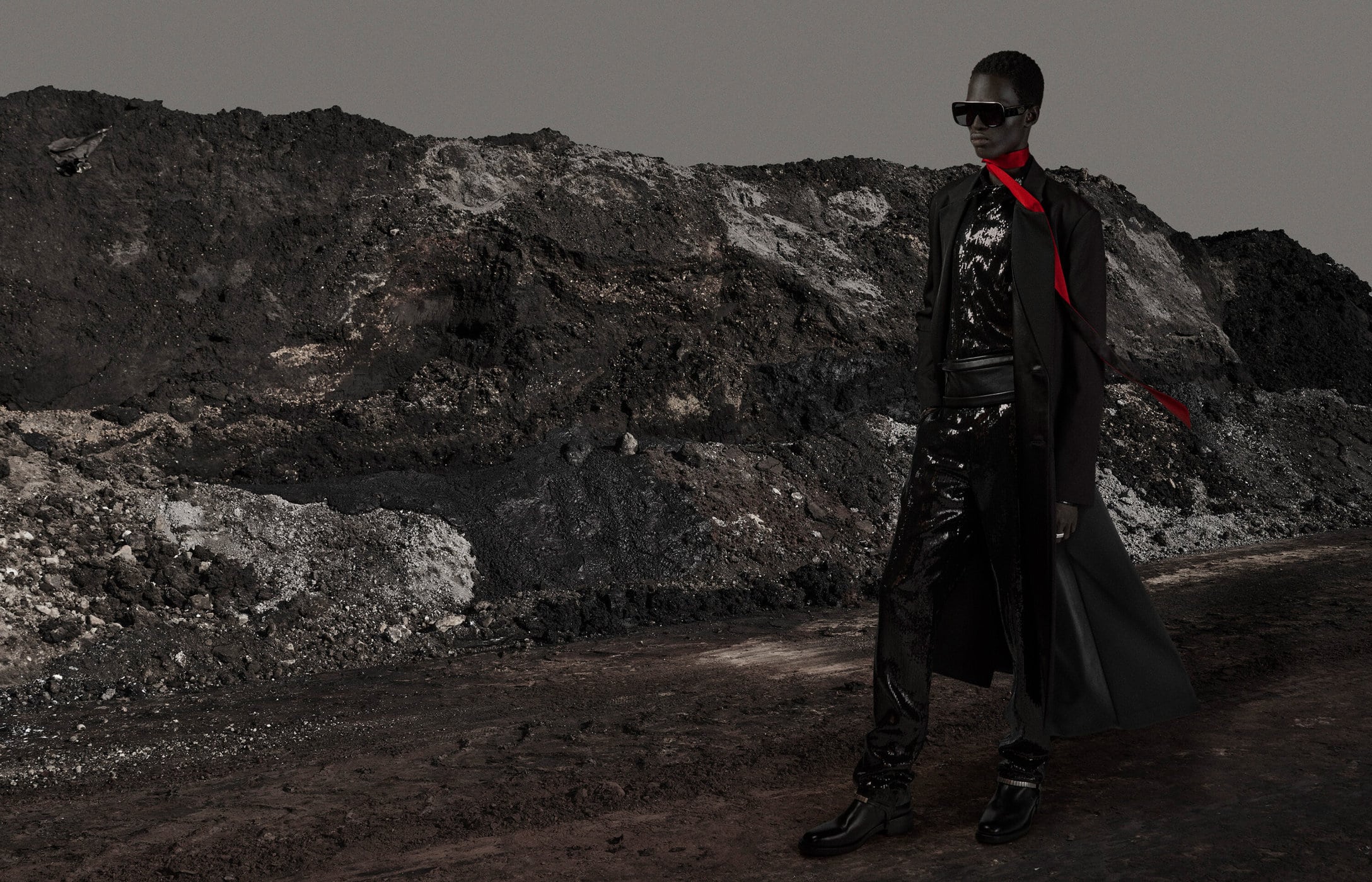 Dark-skinned woman in sunglasses, black coat, and shiny black pants stands on gray dirt, with a dark, textured hill in the background.