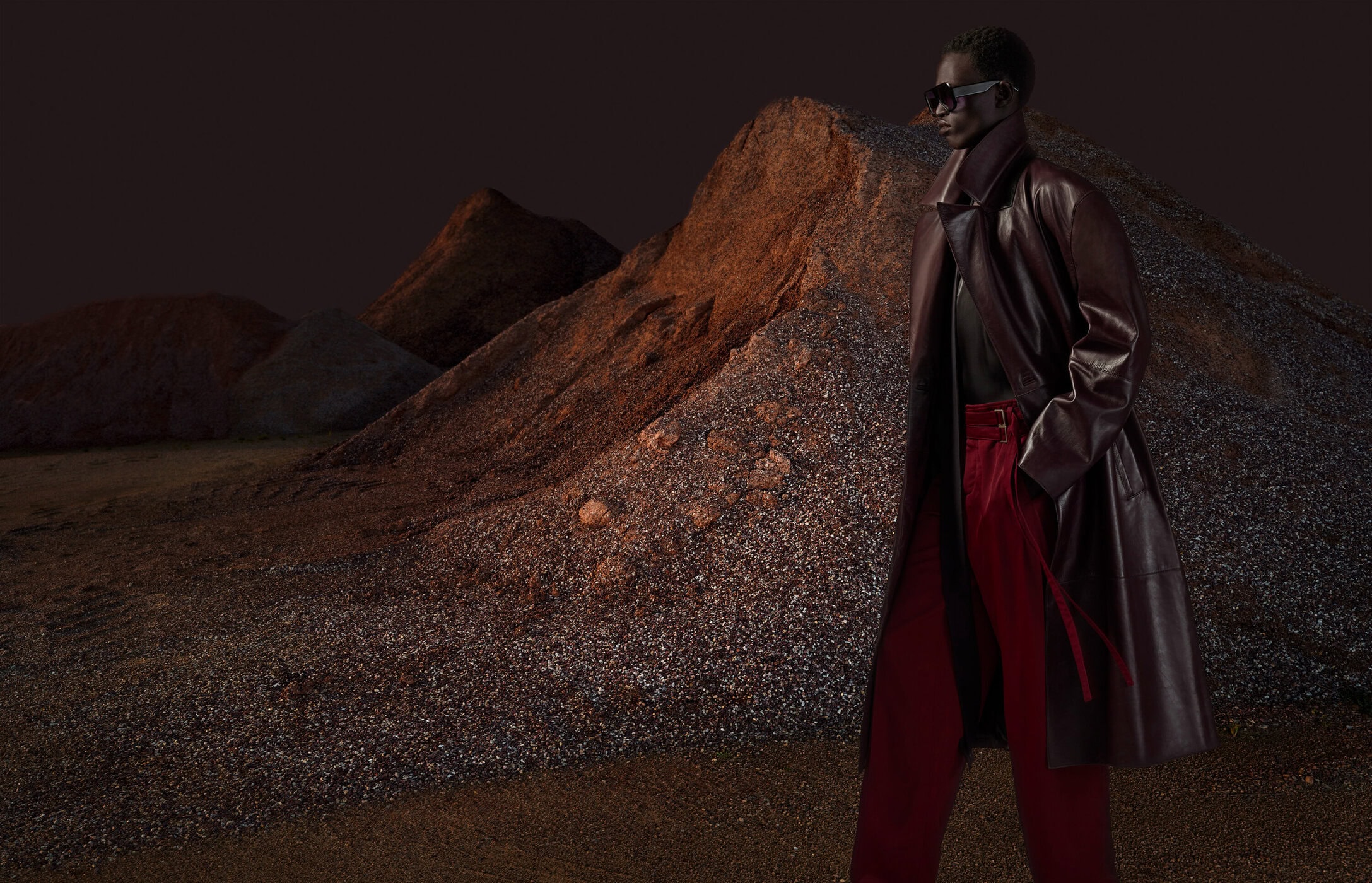Fashion shot of a black model wearing a burgundy coat and red pants in front of heaps of rocks and gravel, dramatic outdoor setting.
