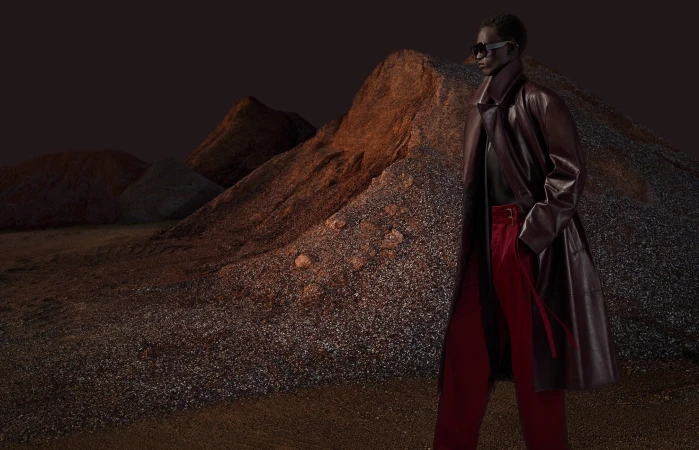 Fashion shot of a black model wearing a burgundy coat and red pants in front of heaps of rocks and gravel, dramatic outdoor setting.