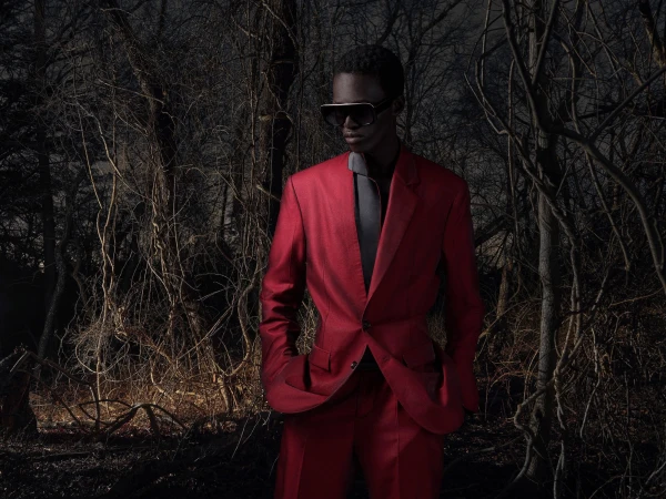 Model wearing a structured red suit and sunglasses standing amidst dark, bare trees in a forest at night