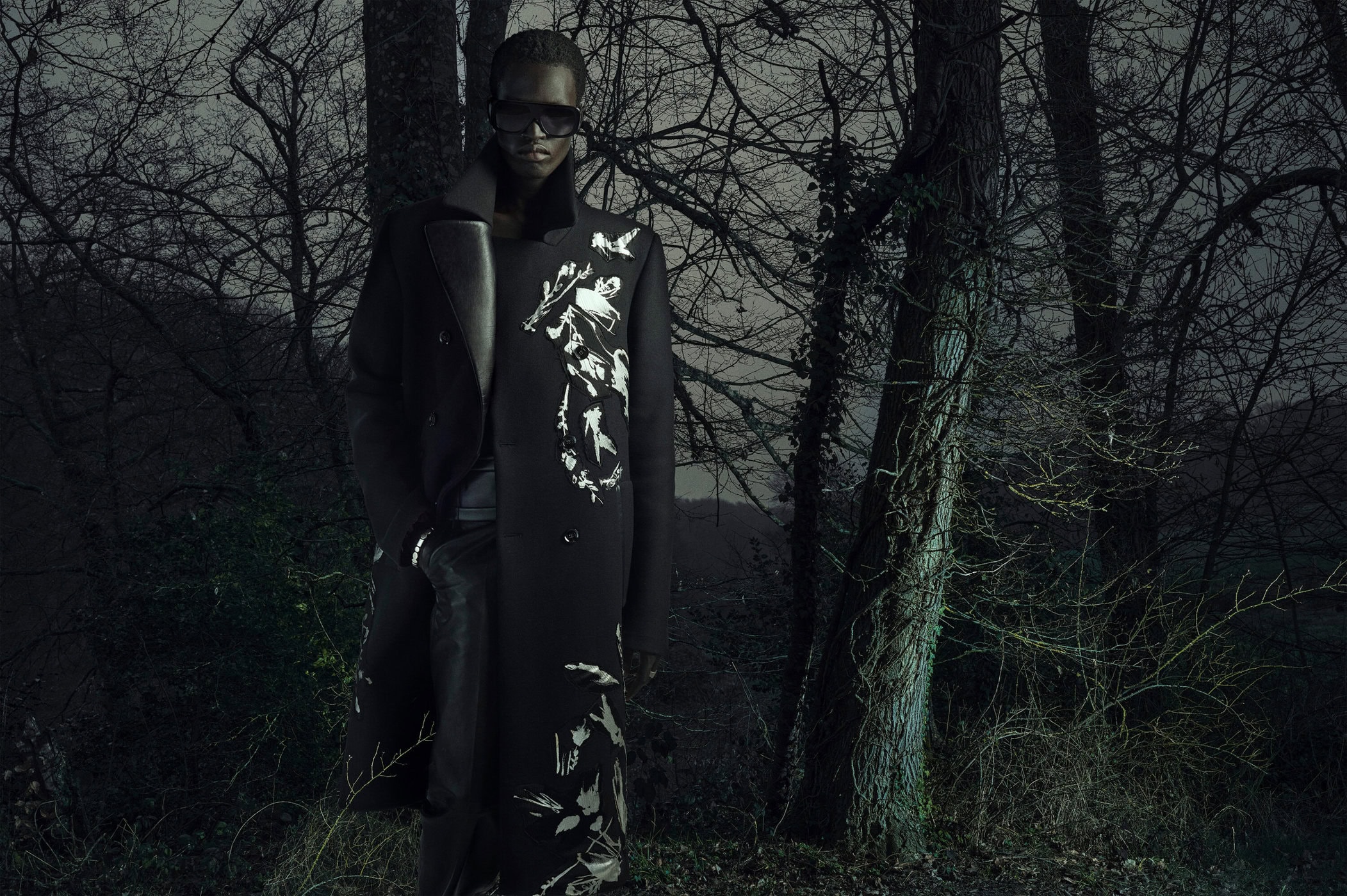 A Black model wearing a long black coat with white abstract patterns, dark sunglasses, and leather pants in a dark, bare winter forest.