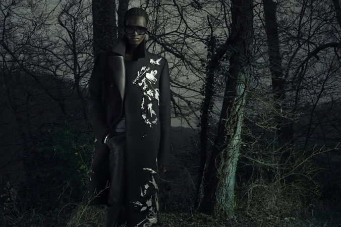A Black model wearing a long black coat with white abstract patterns, dark sunglasses, and leather pants in a dark, bare winter forest.