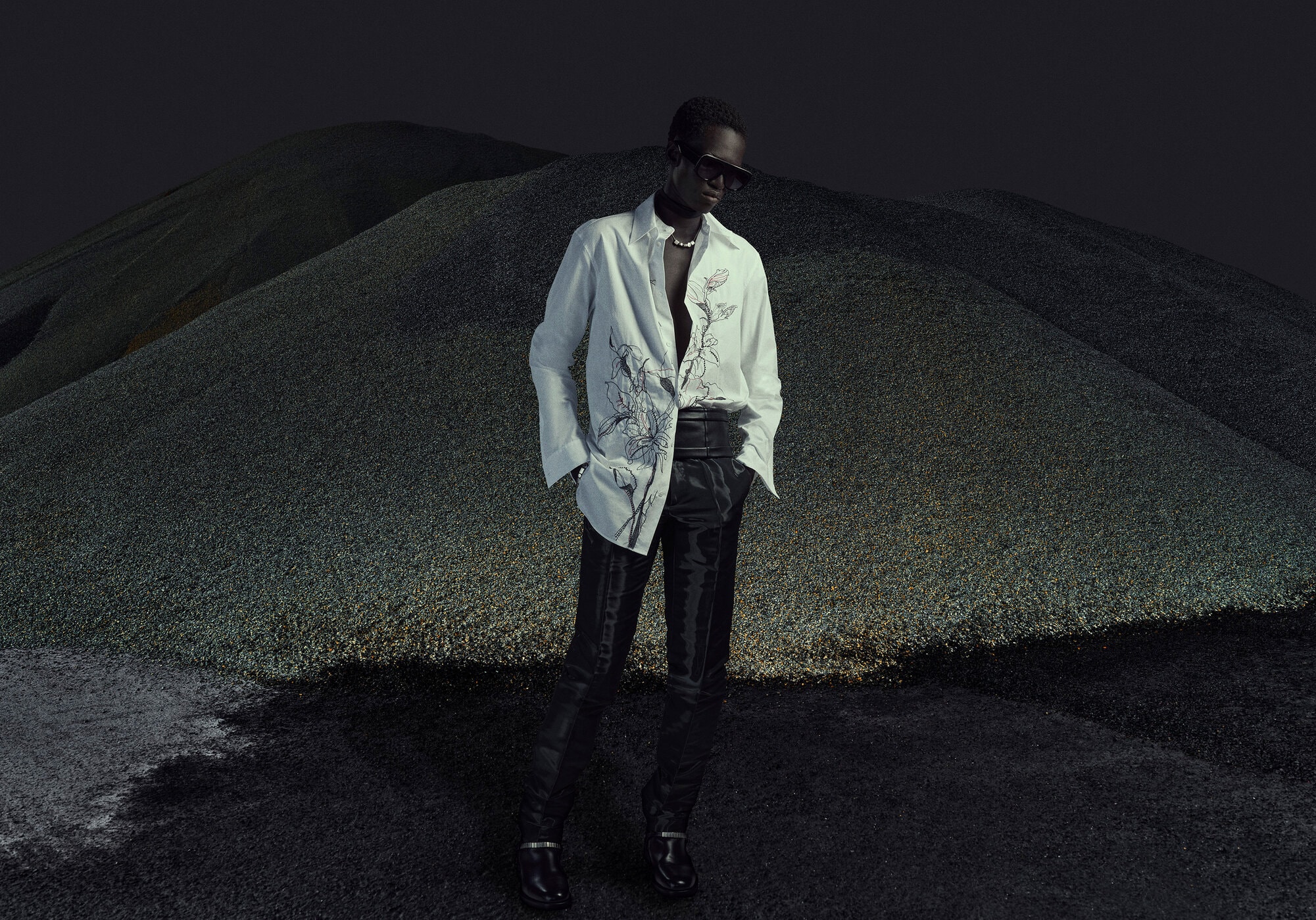 An adult male fashion model poses wearing a patterned white button-down shirt and dark slacks against a dark landscape backdrop.