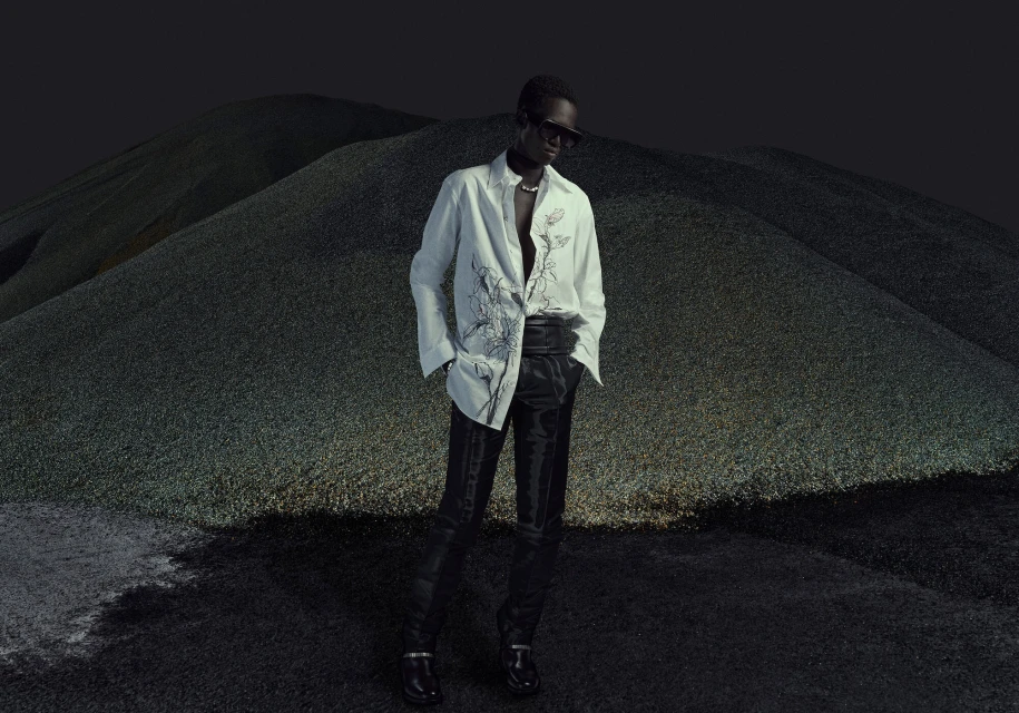 An adult male fashion model poses wearing a patterned white button-down shirt and dark slacks against a dark landscape backdrop.