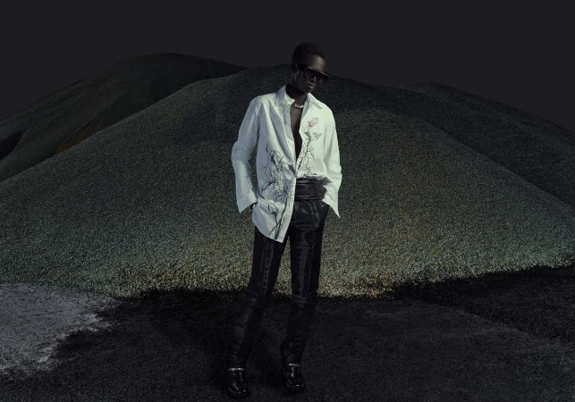 An adult male fashion model poses wearing a patterned white button-down shirt and dark slacks against a dark landscape backdrop.