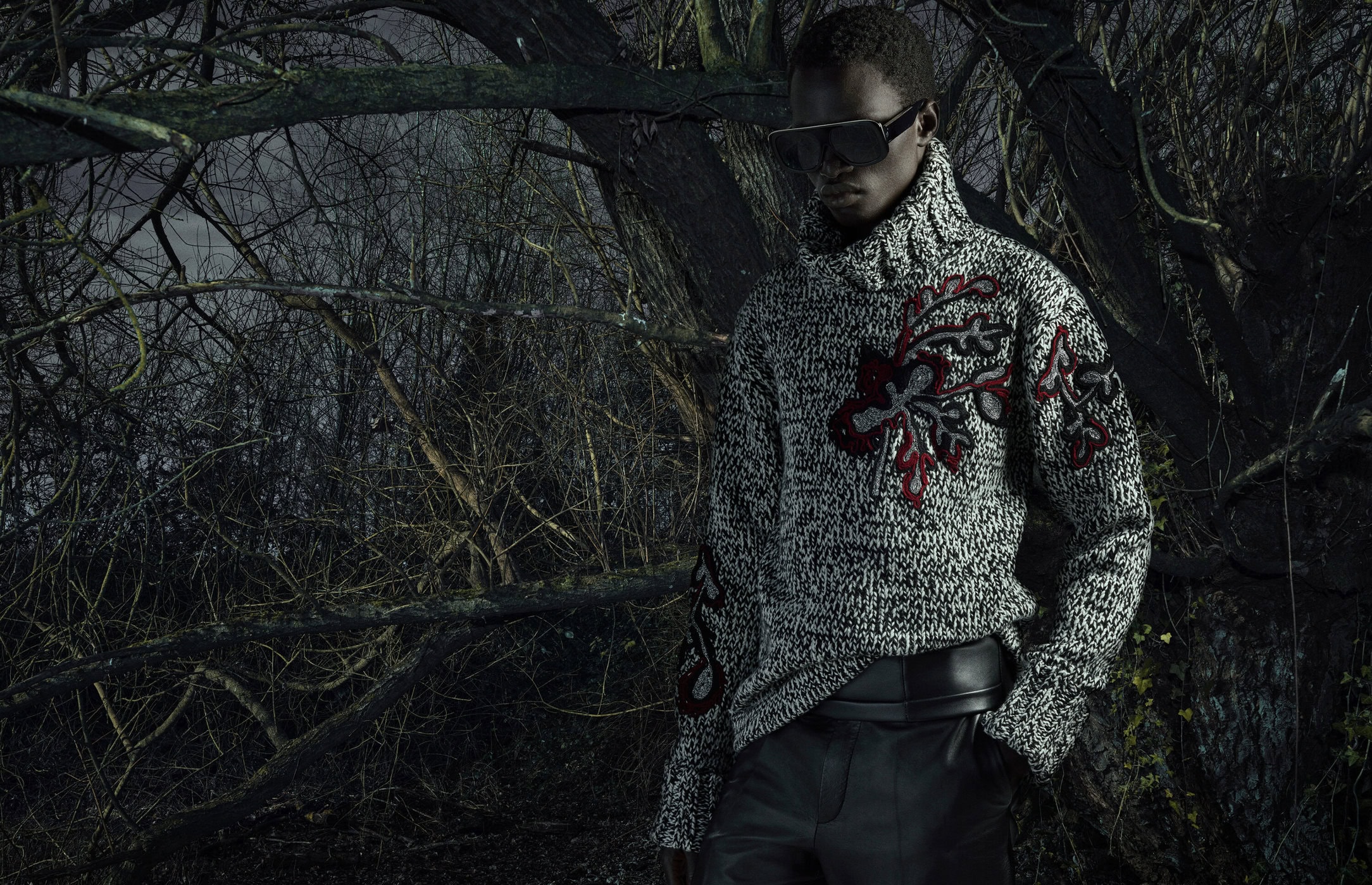 Fashion shot of a Black male model wearing a grey patterned turtleneck sweater and black leather pants in a forest environment.