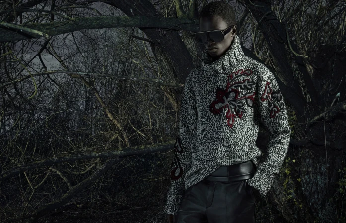 Fashion shot of a Black male model wearing a grey patterned turtleneck sweater and black leather pants in a forest environment.