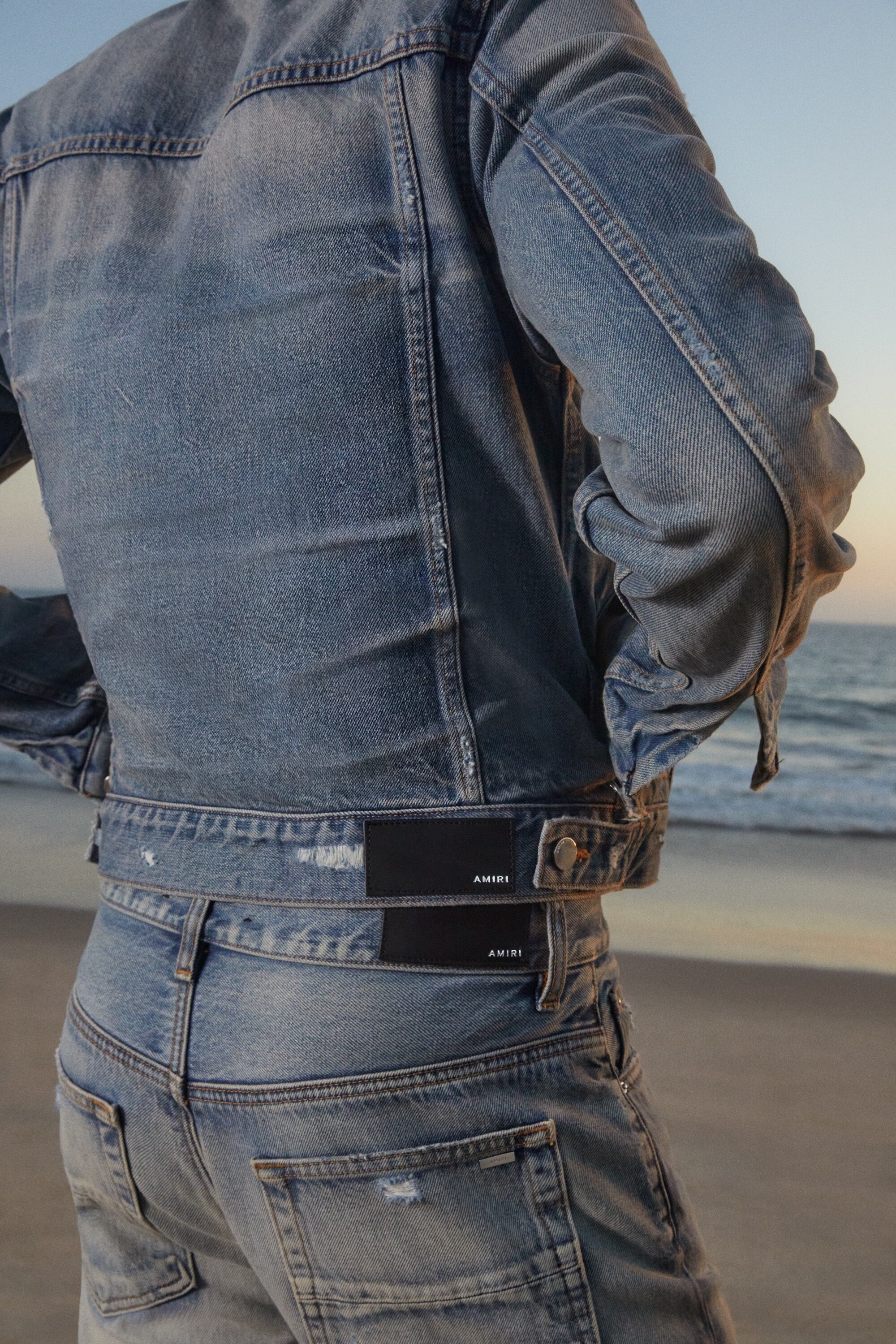 Back view of a person wearing a denim jacket and jeans standing on a beach with the ocean and sky visible in the background.