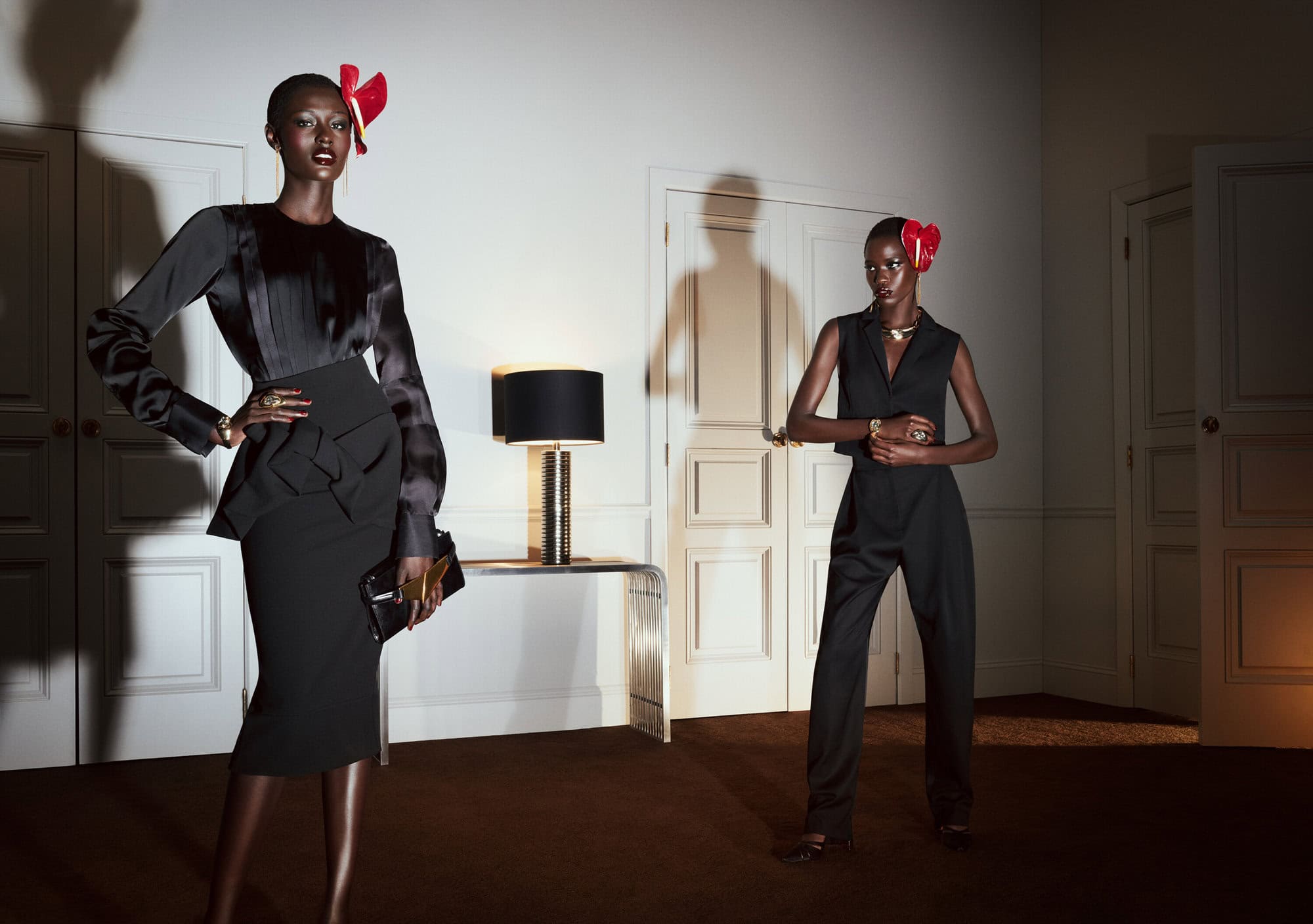 Two Black fashion models pose indoors wearing sophisticated black ensembles with red accessories, complemented by soft, diffused lighting.