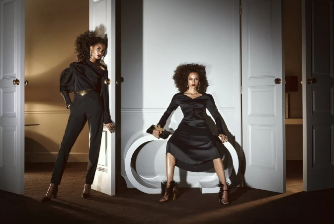 Two African American women in elegant black dresses pose in a doorway with white trim in a fashion-style shot with soft studio lighting.