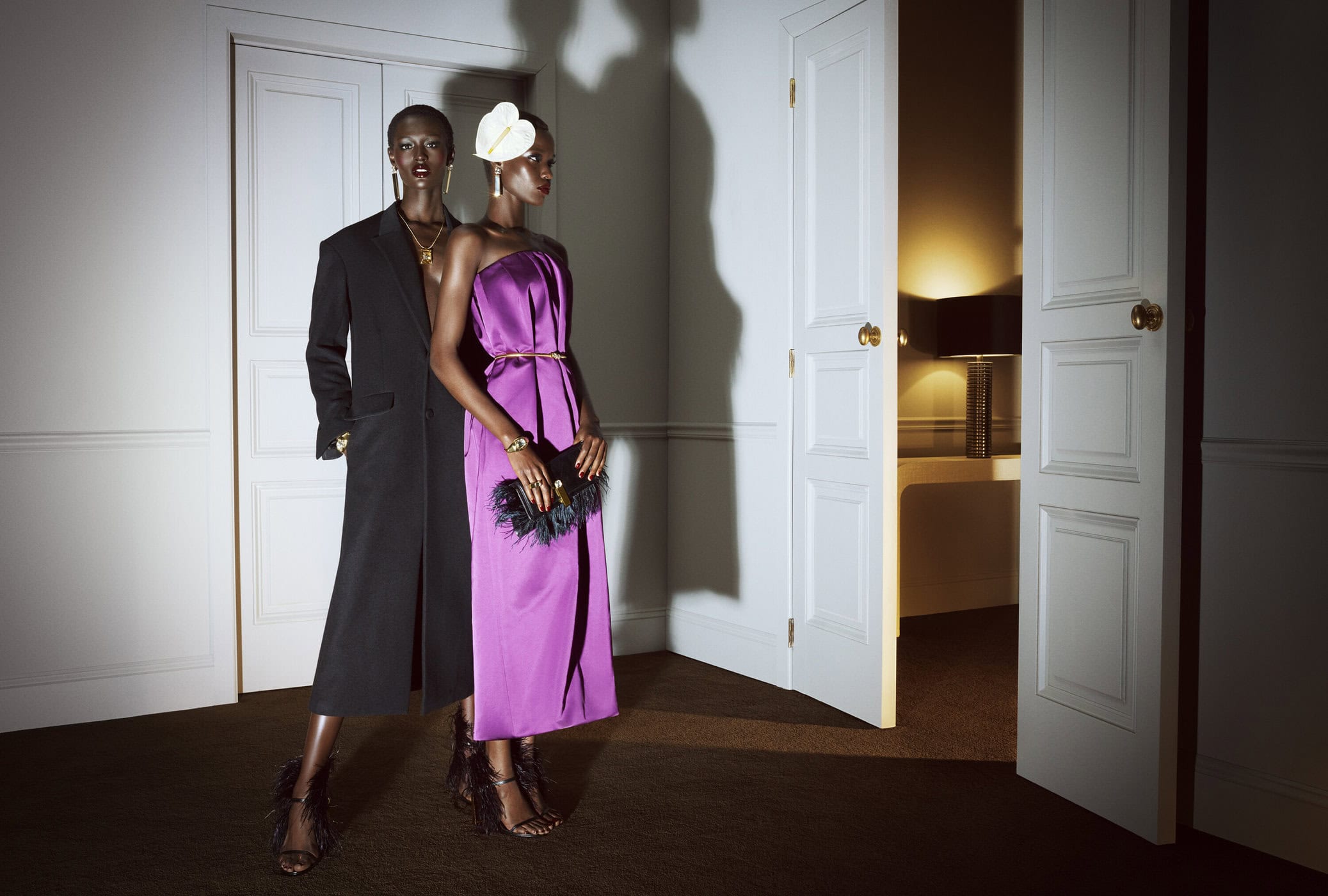 Two black female models in evening wear pose in front of a white door inside a room. One wears a long black coat, the other a purple dress.